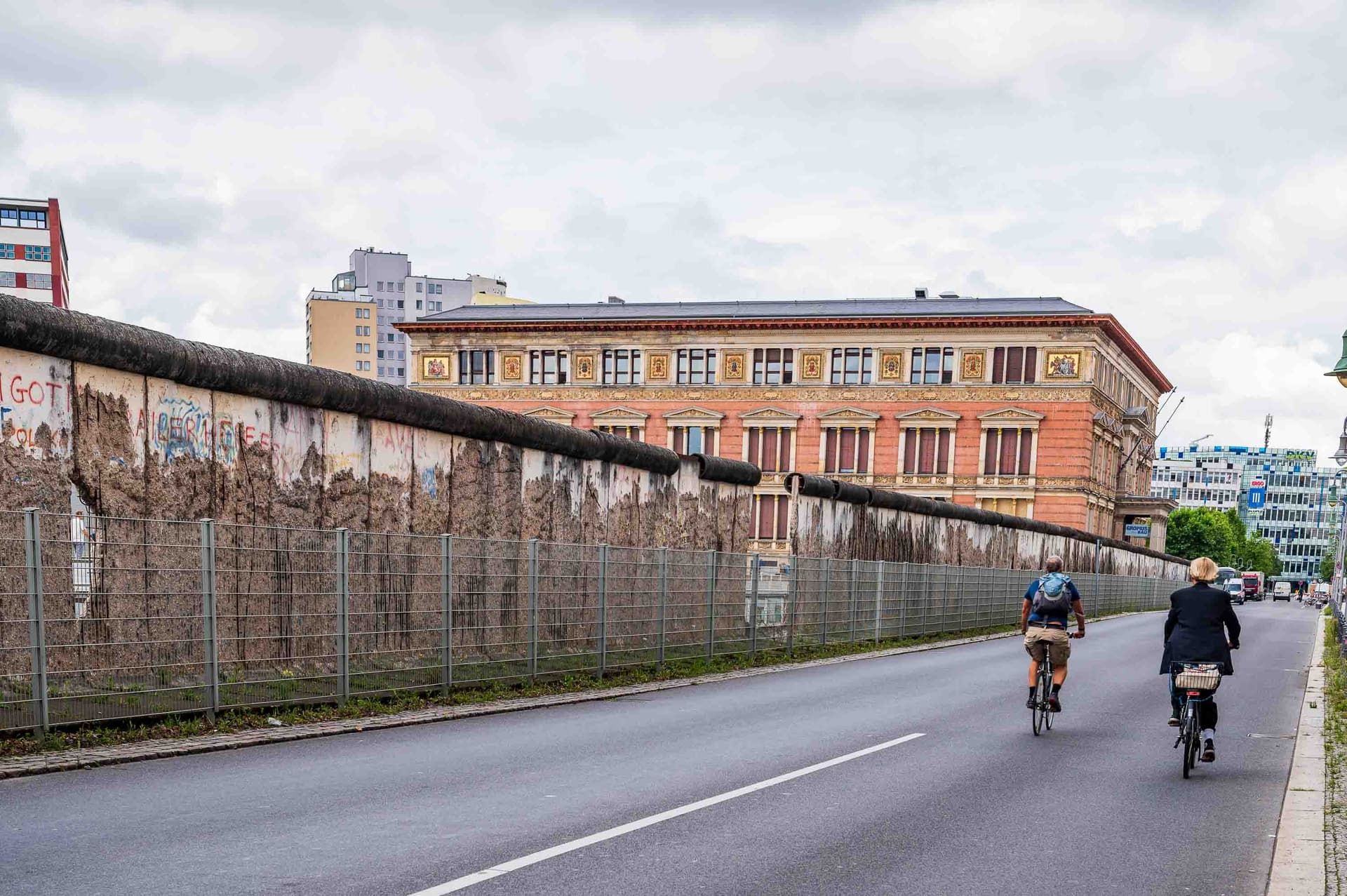 A stretch of the Wall stands beside a historic building while cyclists ride past. History doesn't close; it coexists with the present.