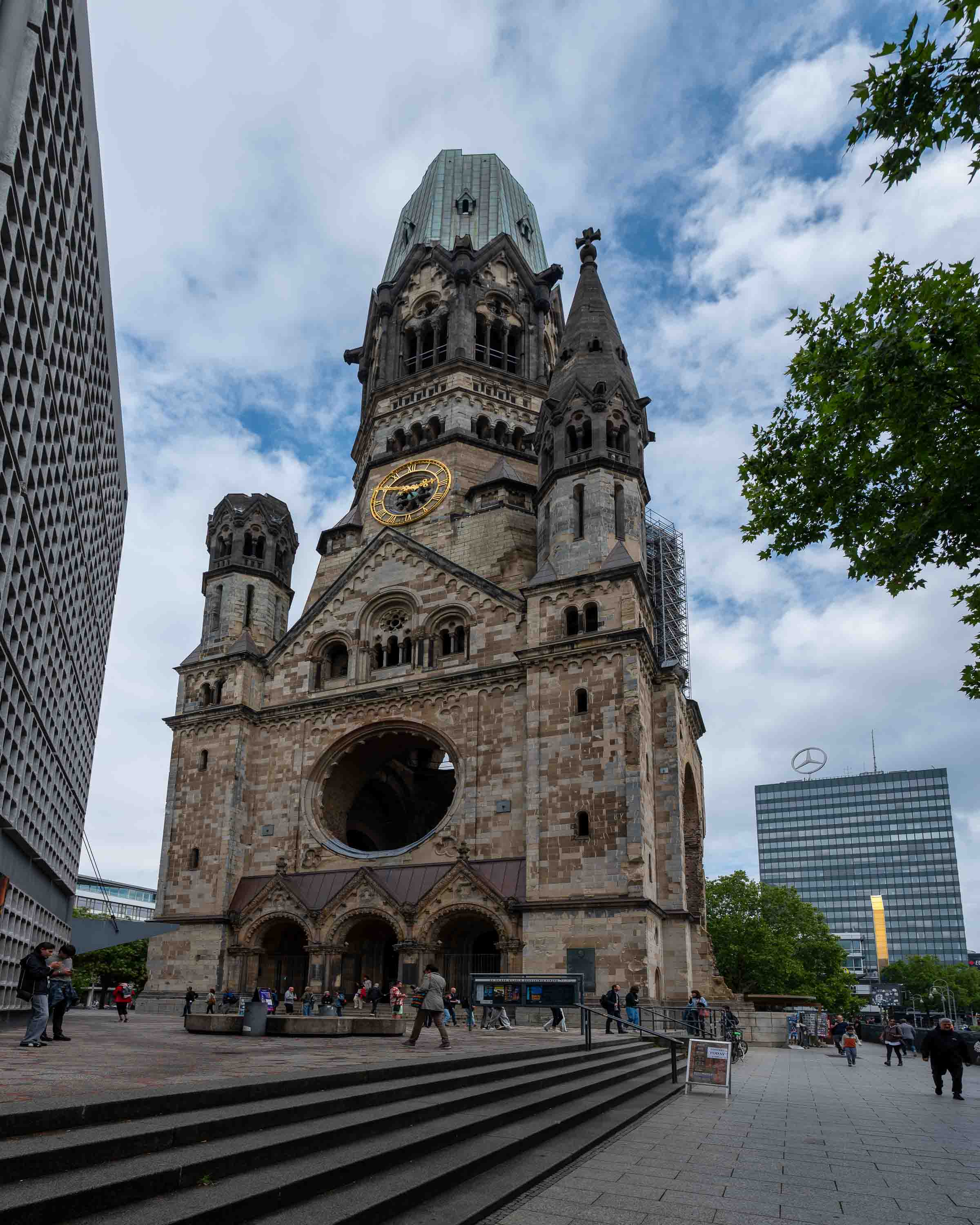 The Kaiser Wilhelm Memorial Church: its tower shattered by Allied bombing in 1943, deliberately preserved as a war memorial. To the right, the Mercedes-Benz logo.