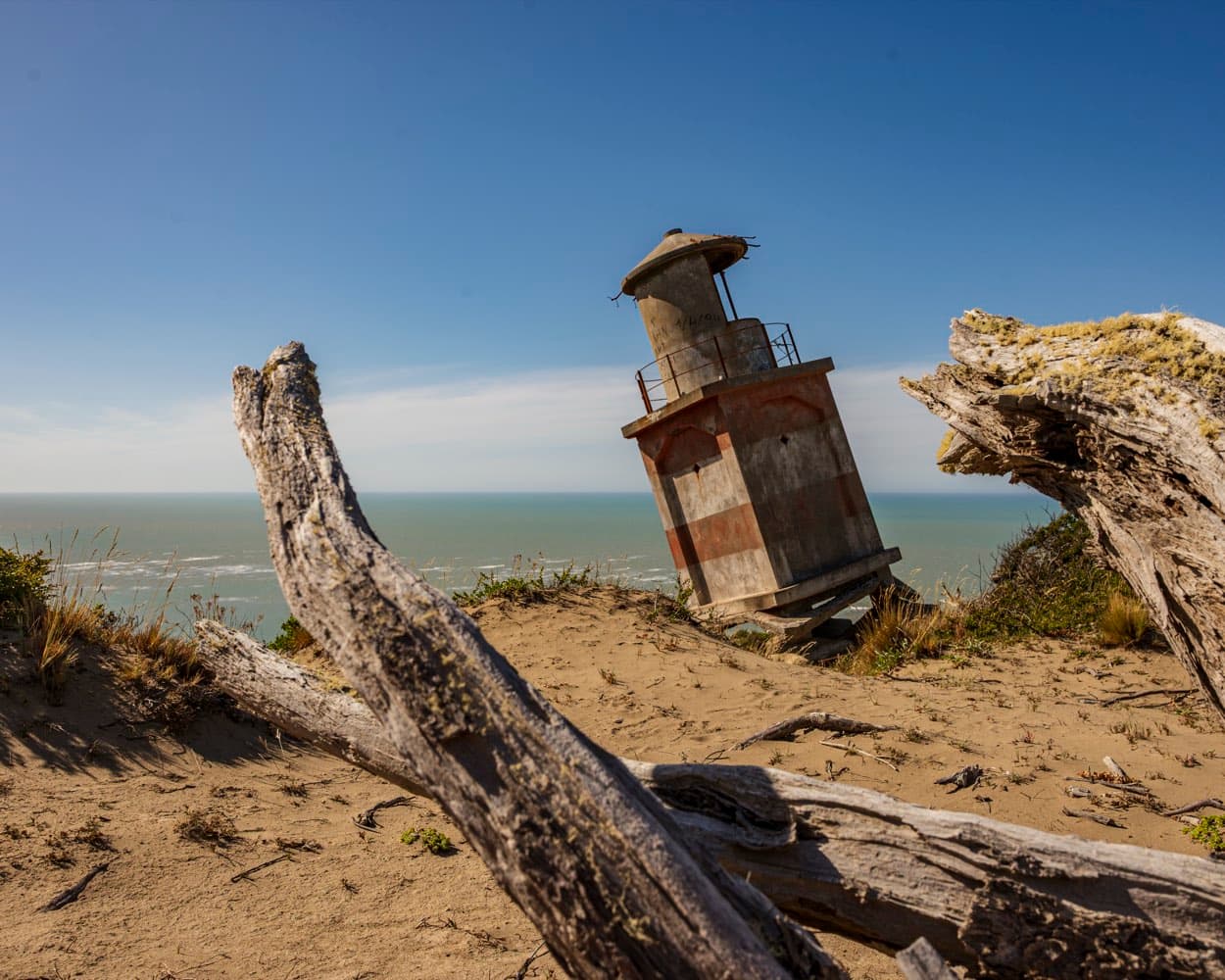 The leaning tower of the original Cabo San Pablo lighthouse among dry logs, with the sea behind